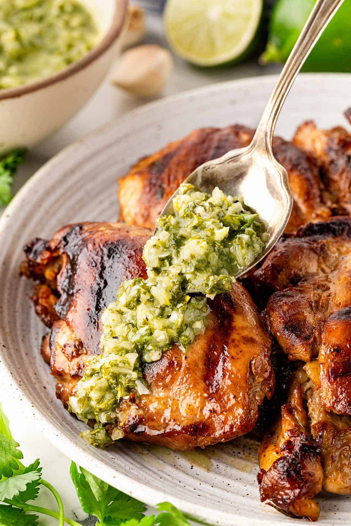 Spoon drizzling chunky chimichurri sauce over grilled chicken thighs on a plate with a blurred kitchen background