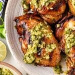 Closeup of grilled chimichurri chicken topped with sauce on a ceramic platter with limes and more sauce in the background