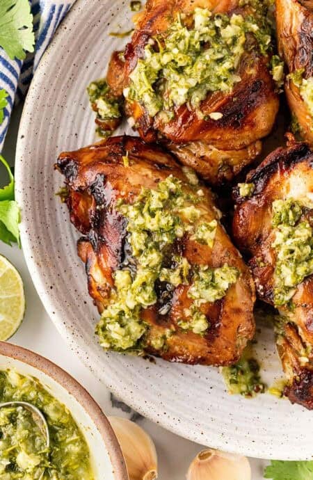 Closeup of grilled chimichurri chicken topped with sauce on a ceramic platter with limes and more sauce in the background