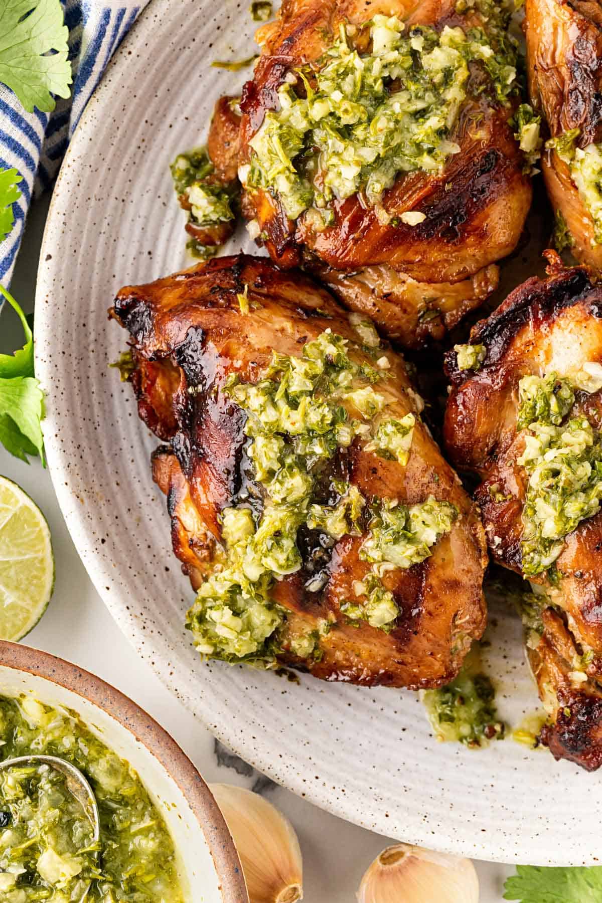 Closeup of grilled chimichurri chicken topped with sauce on a ceramic platter with limes and more sauce in the background