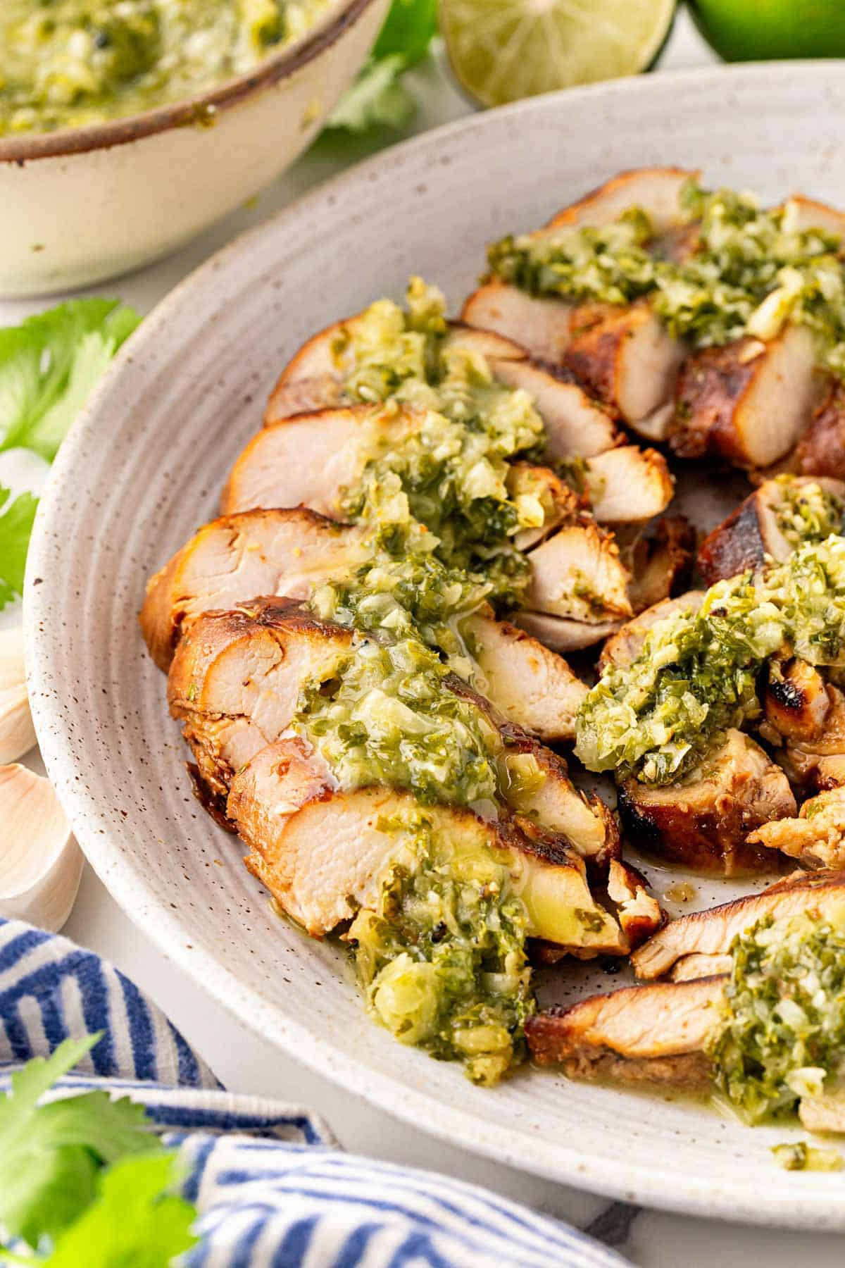 Sliced grilled chimichurri chicken drizzled with sauce on a serving platter with a bowl of sauce and herbs in the background