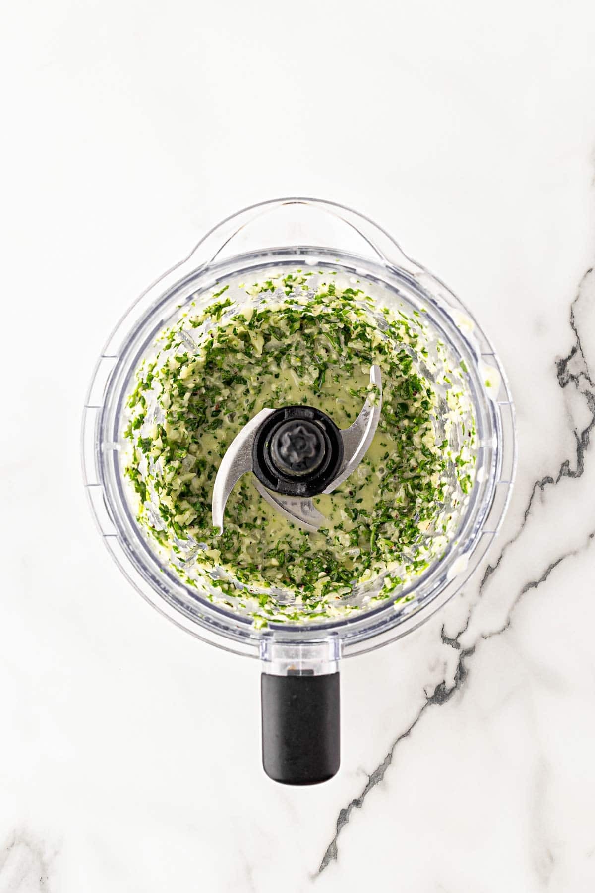 Blended chimichurri sauce in a food processor bowl on a white marble countertop