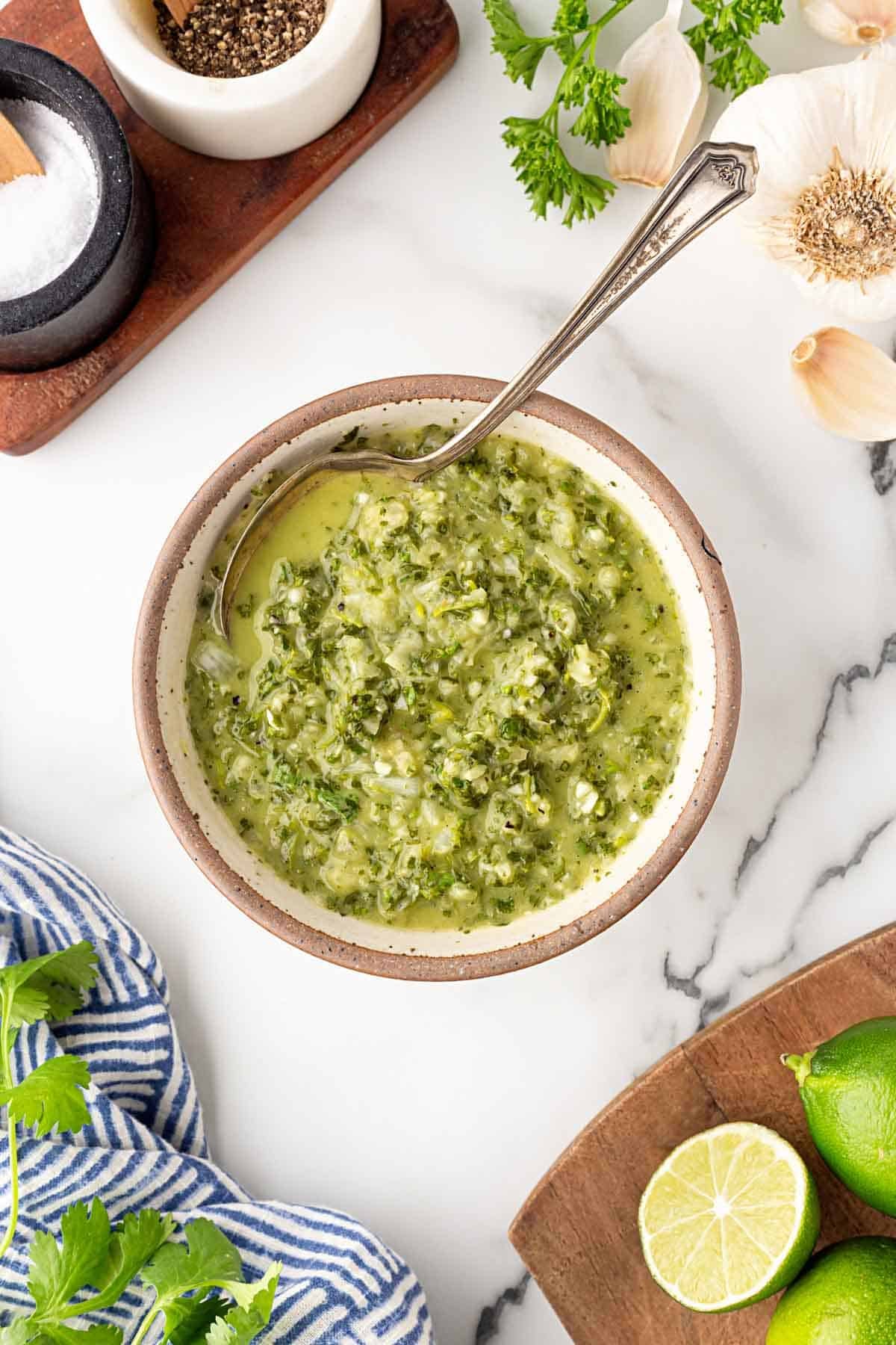 Chimichurri sauce in a bowl with fresh garlic, limes, cilantro, and a striped kitchen towel