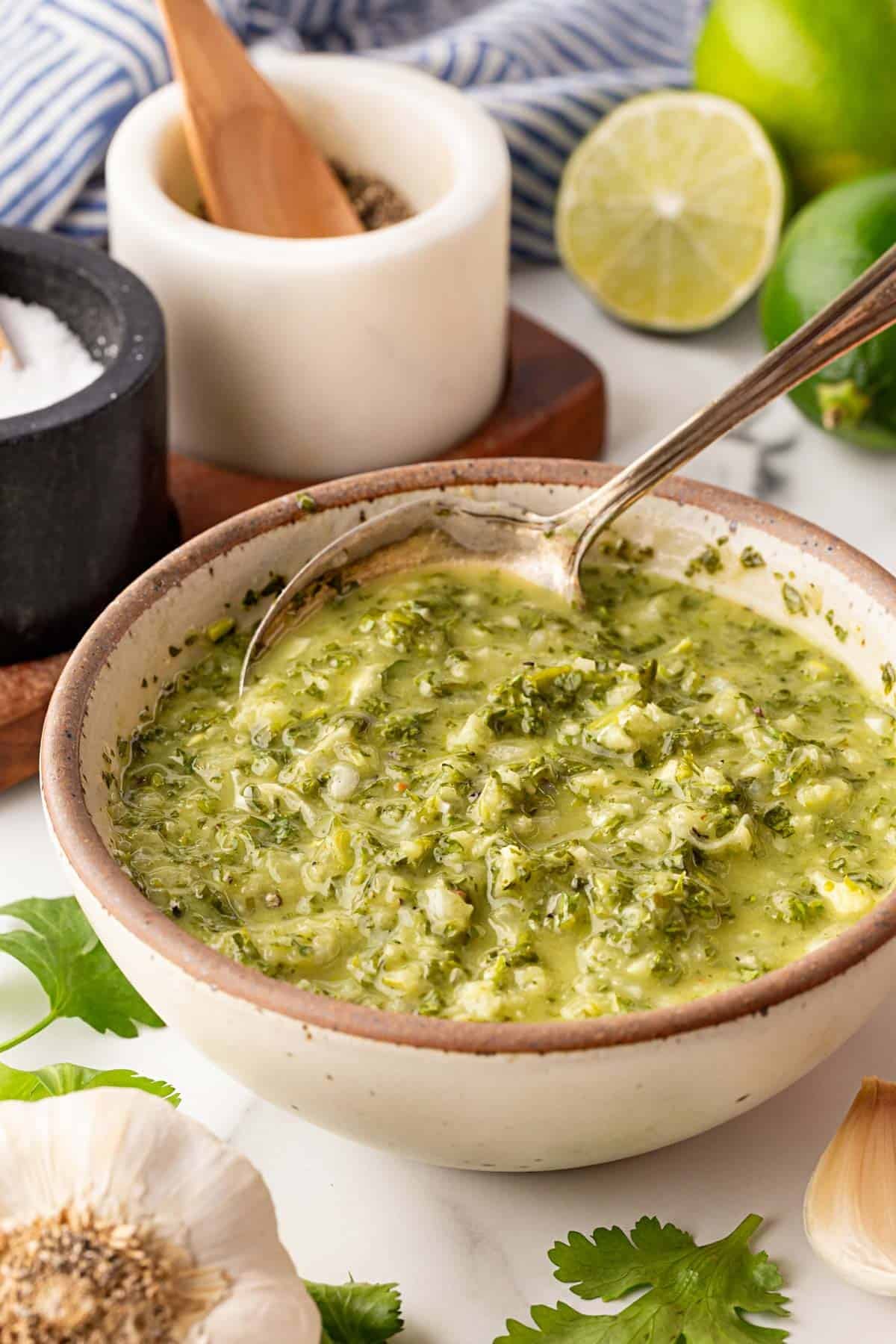 Bowl of homemade chimichurri sauce with a spoon, surrounded by fresh garlic, parsley, and limes on a marble surface