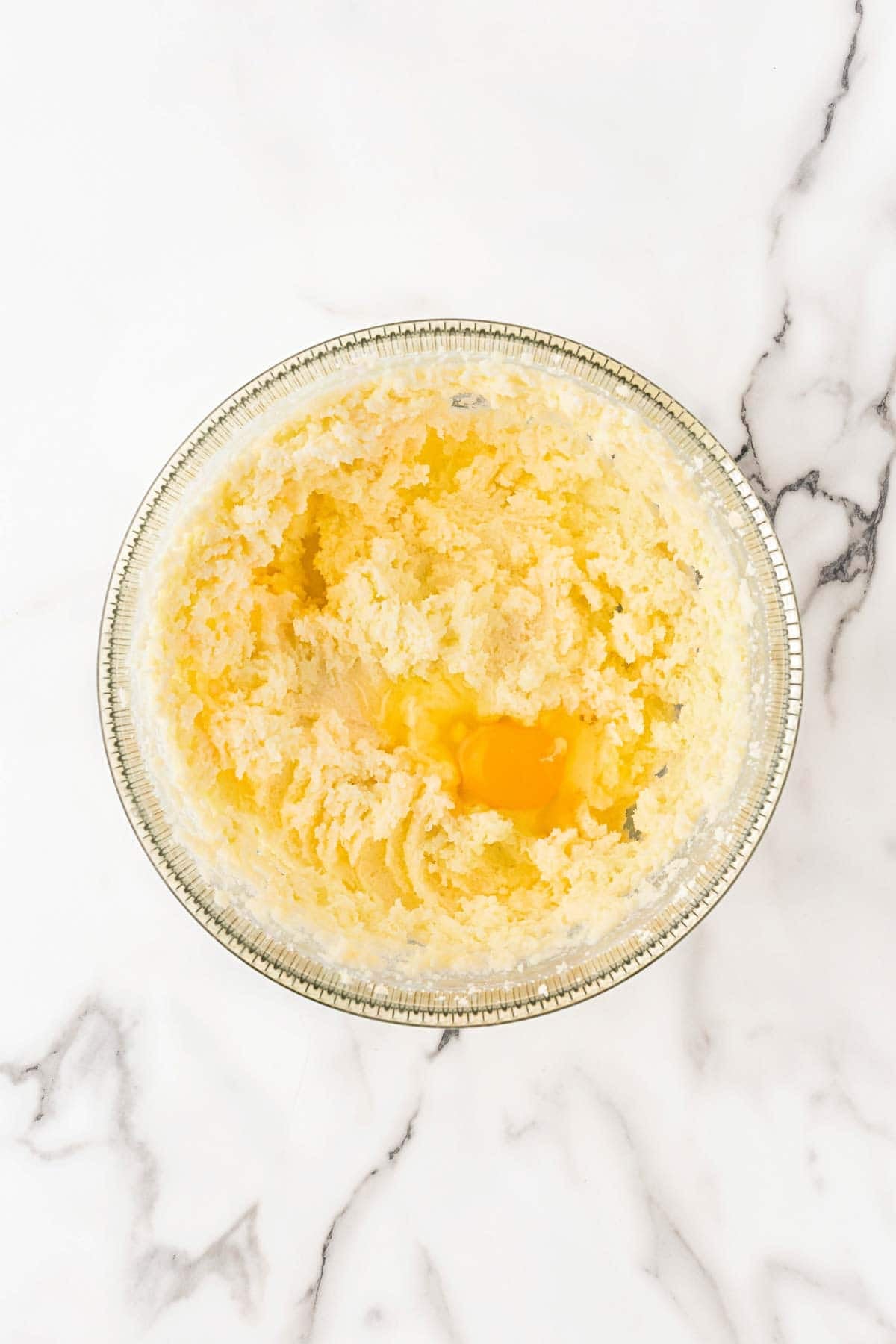 Egg added to a creamed butter and sugar mixture in a clear glass mixing bowl on a marble countertop