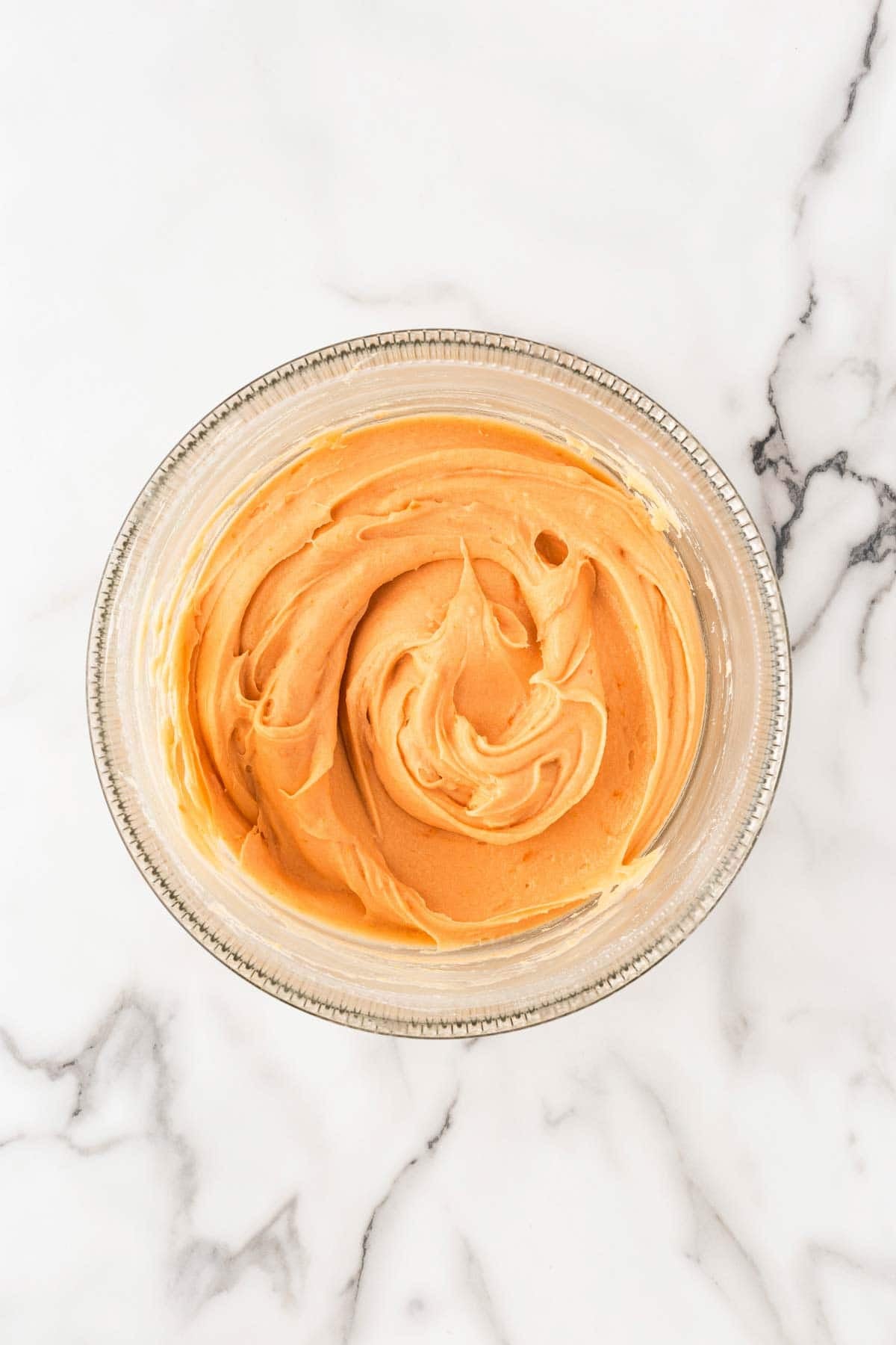 Smooth orange cake batter in a clear glass mixing bowl on a white marble surface