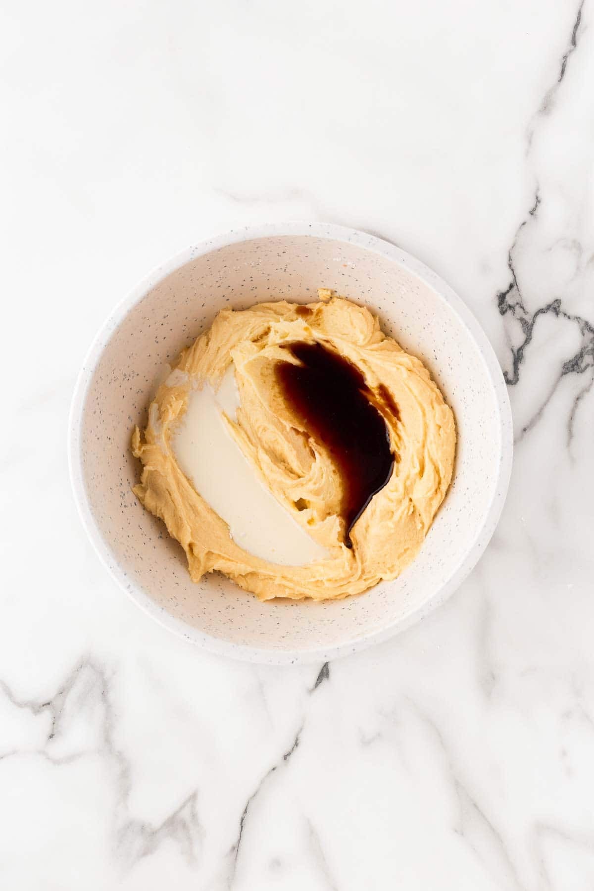 Creamed butter mixture with vanilla extract and heavy cream added in a white mixing bowl on a marble countertop