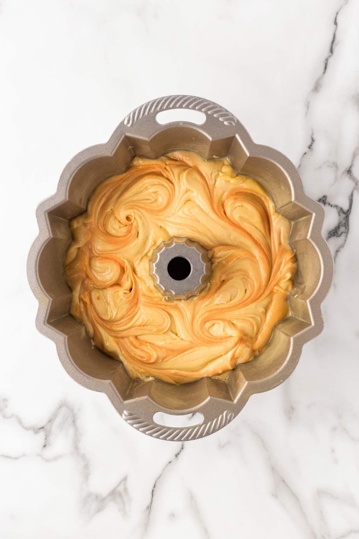 Orange and vanilla cake batter swirled together in a fluted Bundt pan on a marble countertop before baking