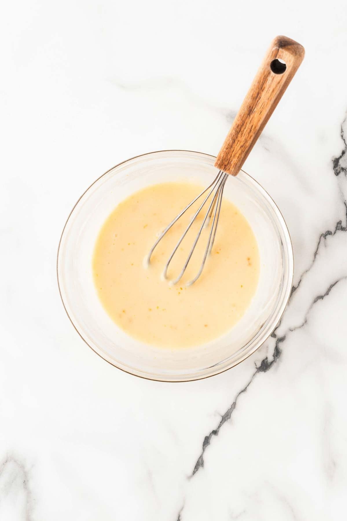 Orange glaze whisked until smooth in a clear glass bowl with a wooden-handled whisk on a marble countertop