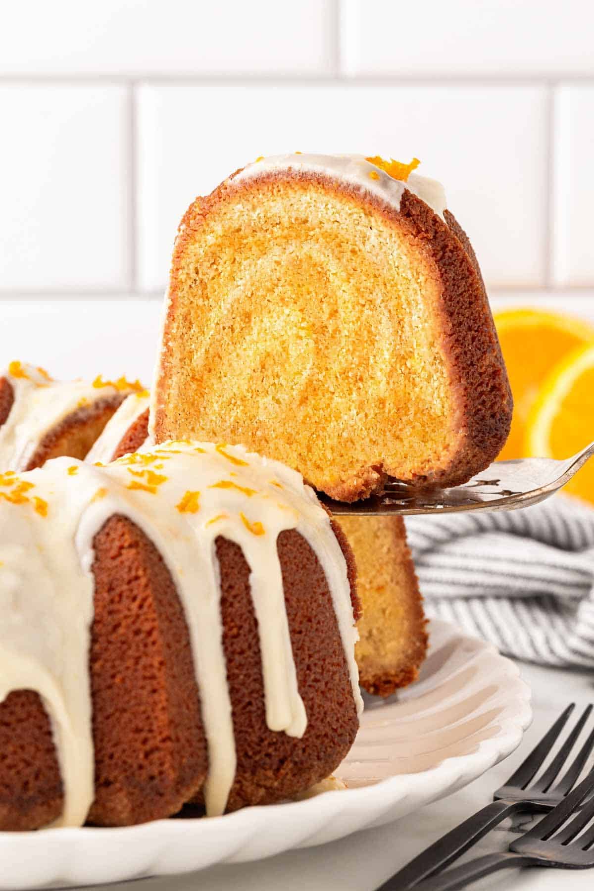 Thick slice of creamsicle pound cake lifted on a cake server with the glazed Bundt cake and orange slices in the background