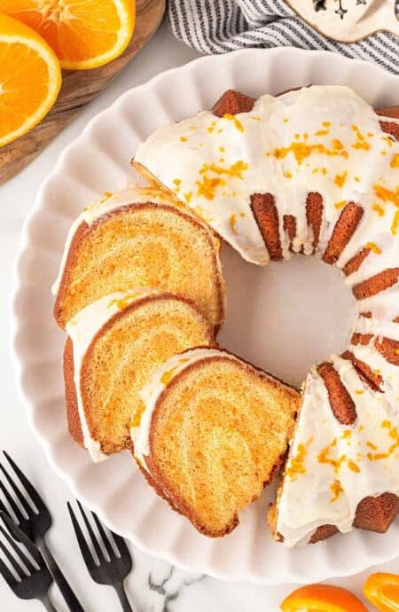 Overhead shot of sliced creamsicle pound cake with orange glaze and fresh zest on a white serving platter with orange halves nearby