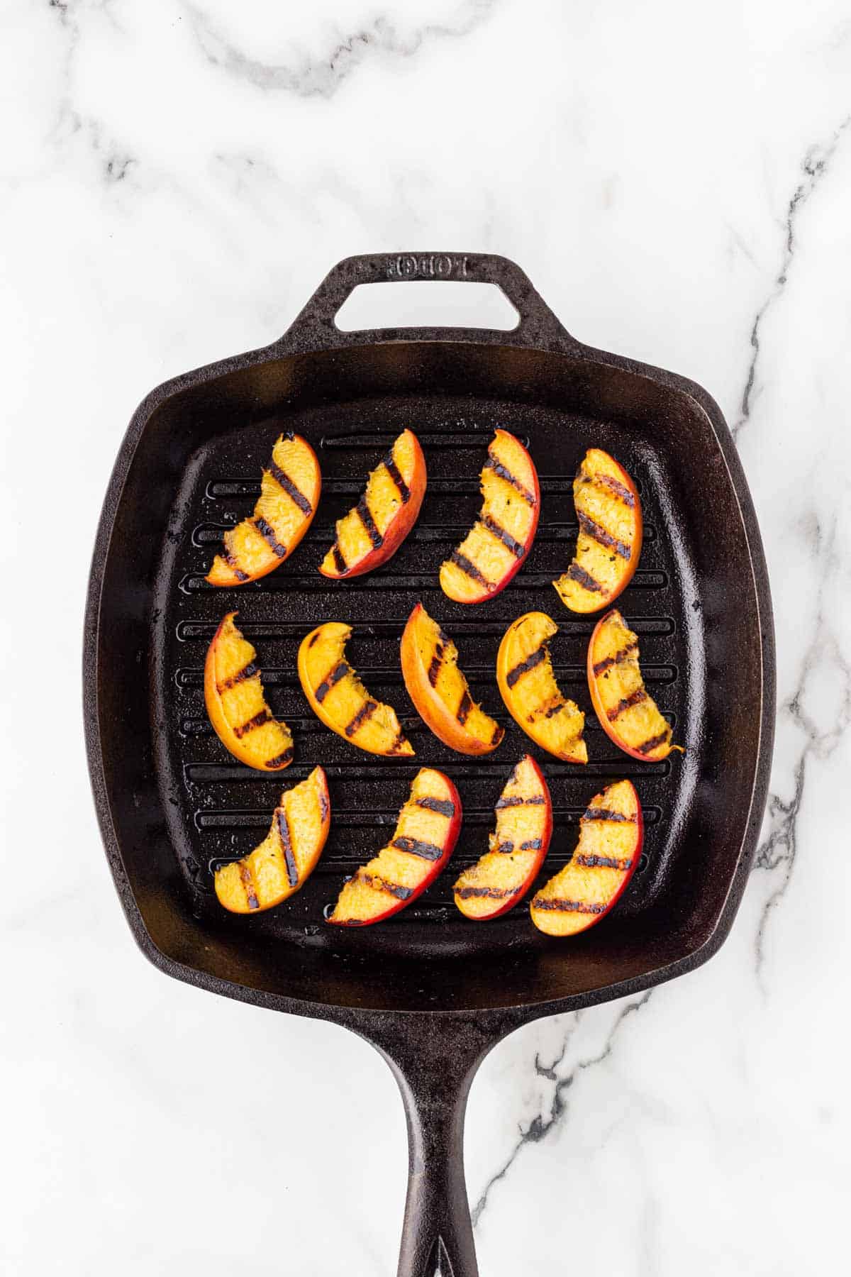 Grilled peach wedges with char marks arranged in a black cast iron grill pan on a white marble surface