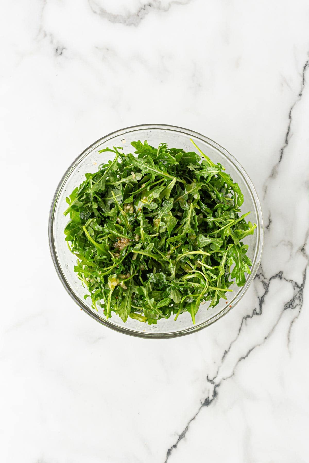 Arugula tossed with citrus dressing in a clear glass bowl on a white marble countertop