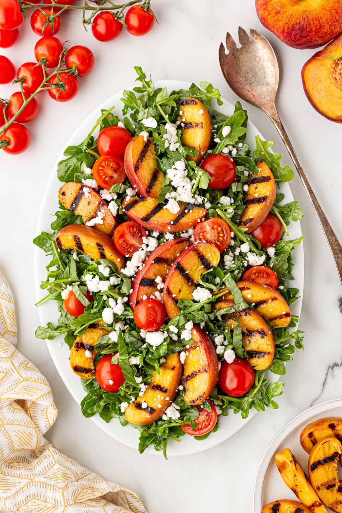 Overhead shot of grilled peach salad with arugula, cherry tomatoes, goat cheese, and basil on a white platter over a light marble surface