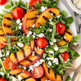 Overhead shot of grilled peach salad with arugula, cherry tomatoes, crumbled goat cheese, and basil, surrounded by fresh peaches and tomatoes on a marble countertop