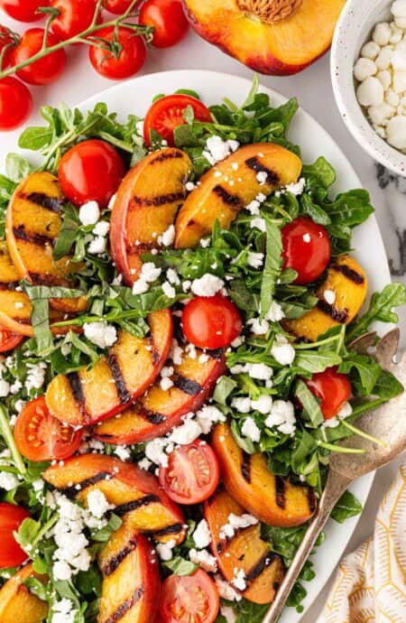 Overhead shot of grilled peach salad with arugula, cherry tomatoes, crumbled goat cheese, and basil, surrounded by fresh peaches and tomatoes on a marble countertop