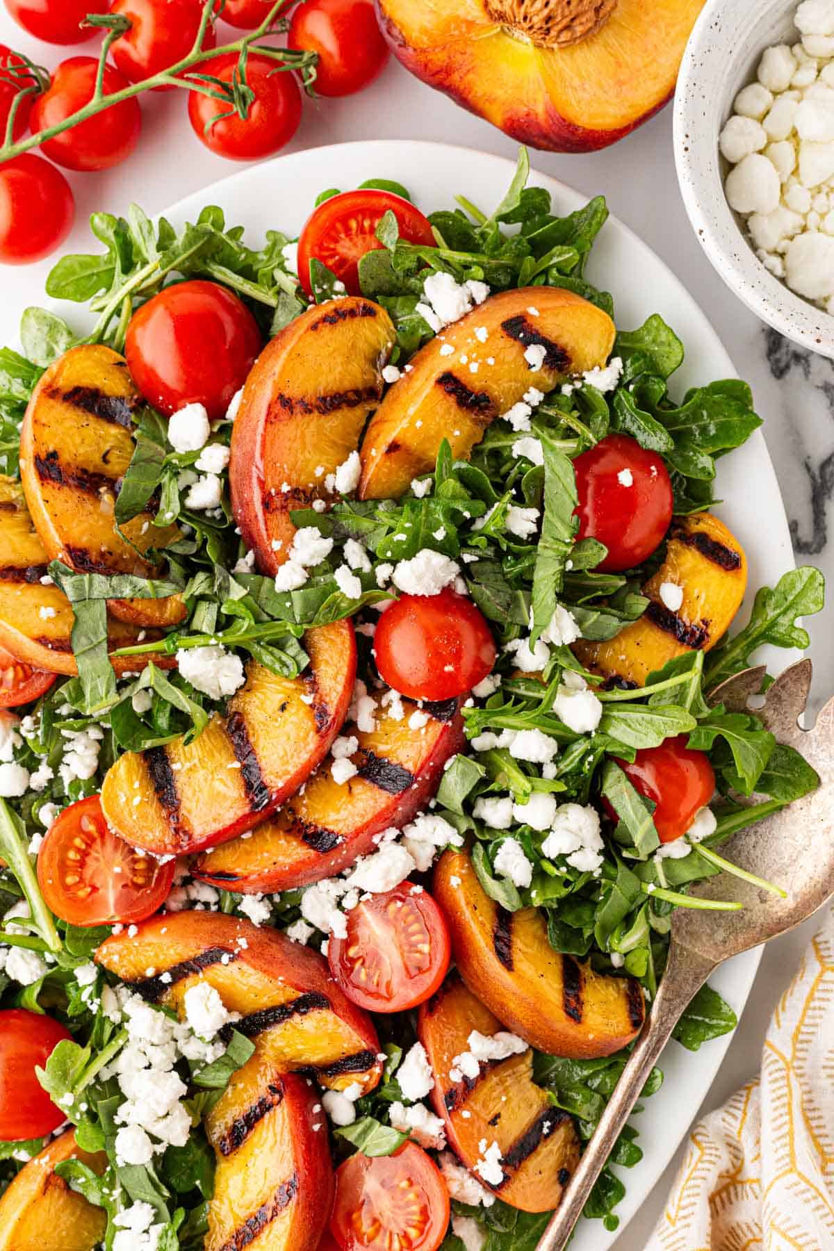 Overhead shot of grilled peach salad with arugula, cherry tomatoes, crumbled goat cheese, and basil, surrounded by fresh peaches and tomatoes on a marble countertop
