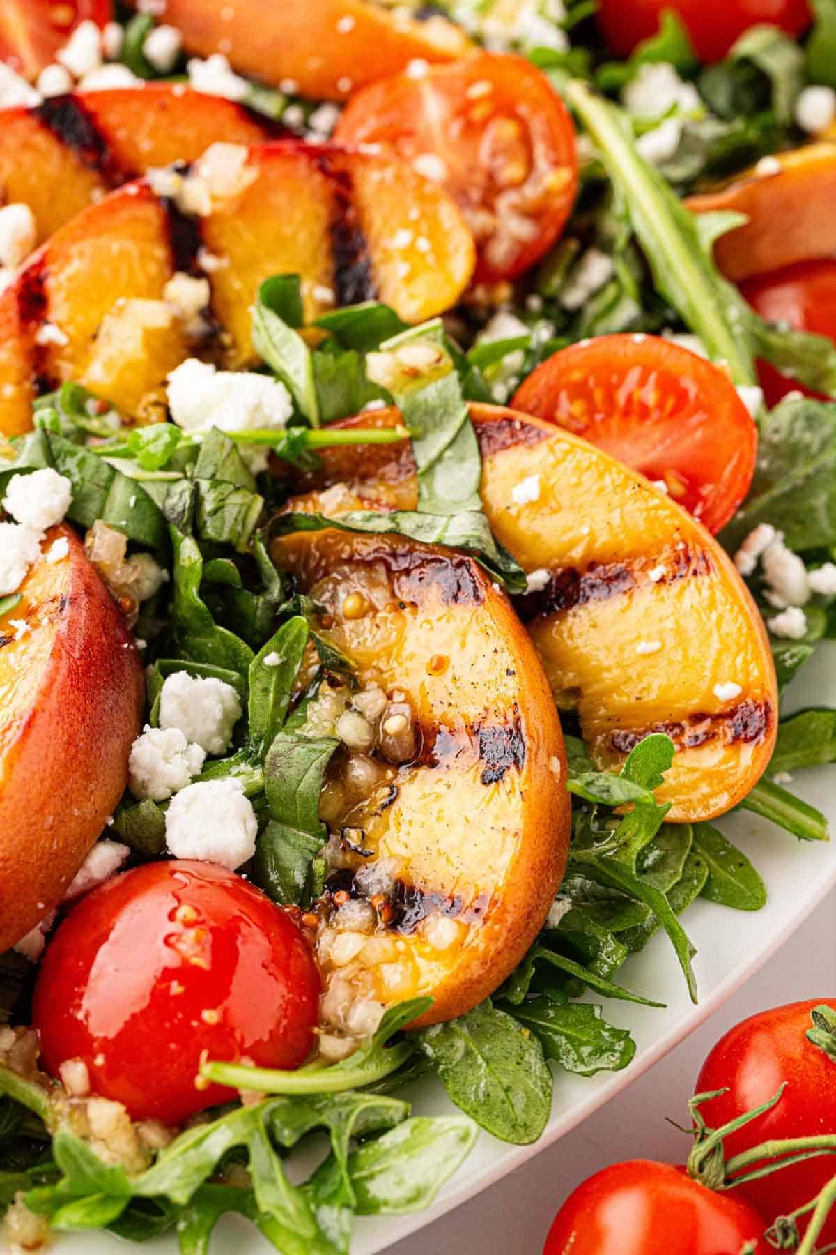 Closeup of grilled peach salad drizzled with citrus dressing, highlighting charred peaches, basil, cherry tomatoes, and goat cheese on a white platter
