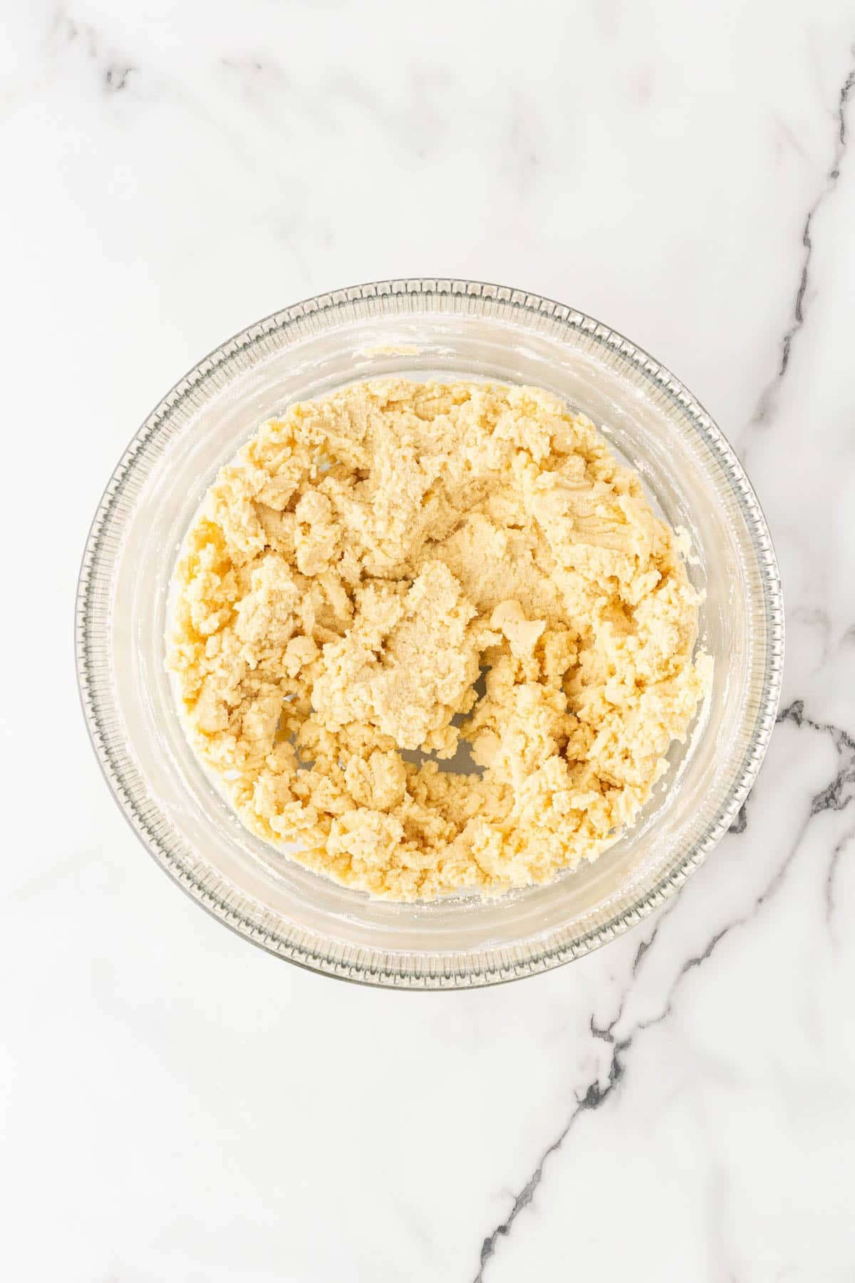 Lemon bar crust mixture with a crumbly texture in a clear glass mixing bowl on a marble countertop
