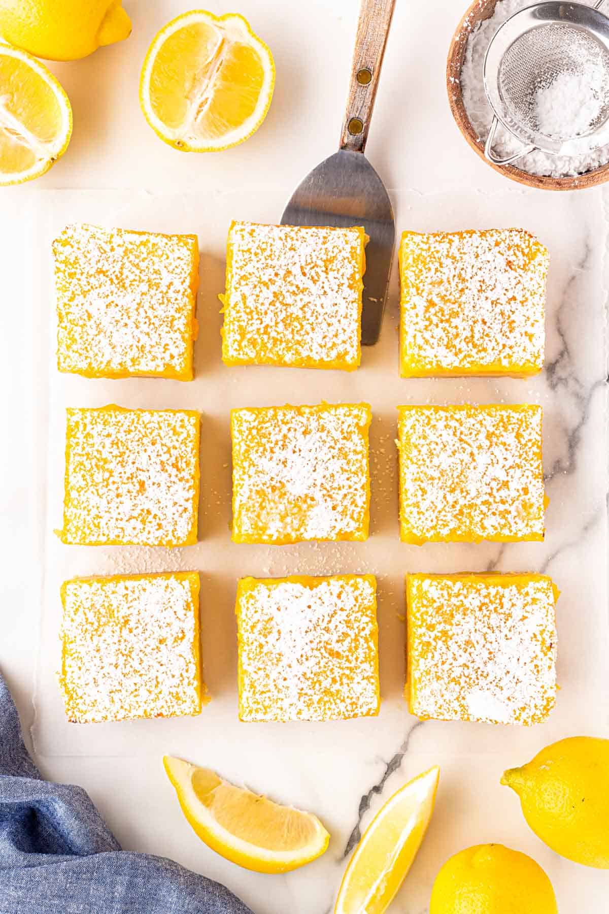 Overhead shot of lemon bars dusted with powdered sugar, arranged on parchment paper with fresh lemons, a spatula, and a small sieve of powdered sugar