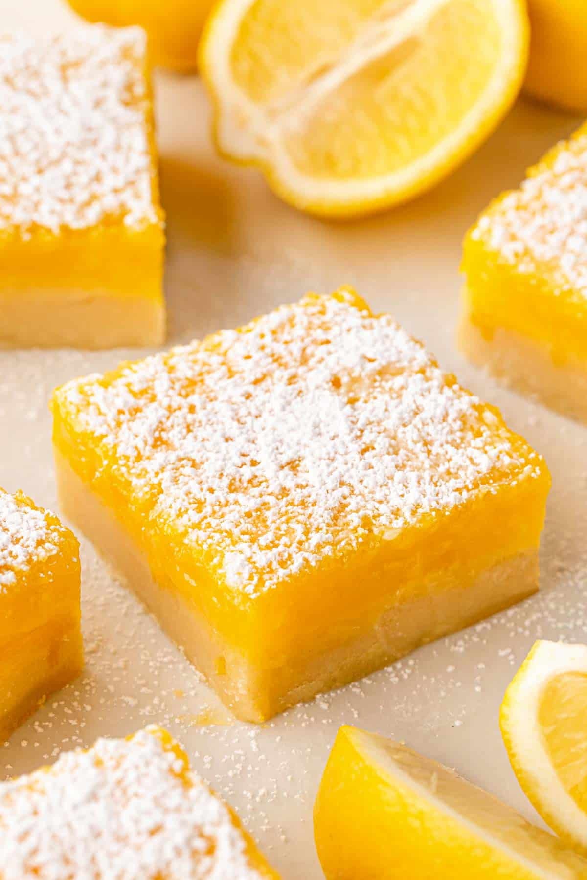 Lemon bar dusted with powdered sugar on parchment paper, with lemon slices and more bars blurred in the background