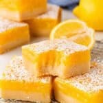 Lemon bars on a cooling rack, including one with a bite taken out, with lemons and a blue towel in the background