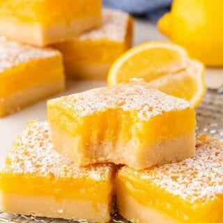 Lemon bars on a cooling rack, including one with a bite taken out, with lemons and a blue towel in the background