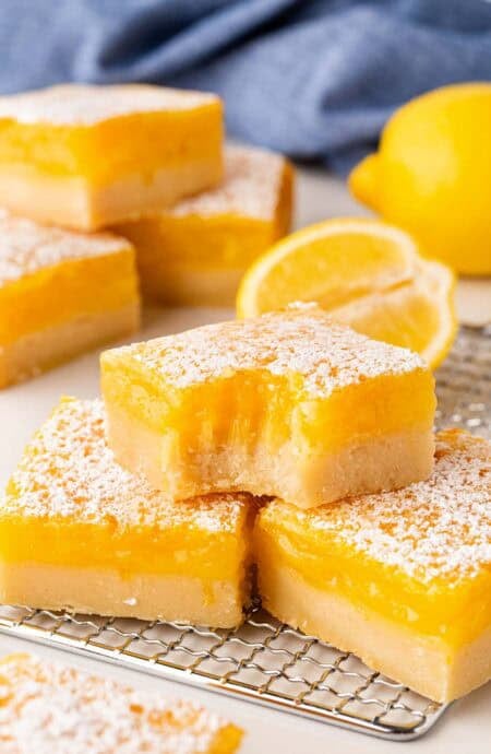 Lemon bars on a cooling rack, including one with a bite taken out, with lemons and a blue towel in the background