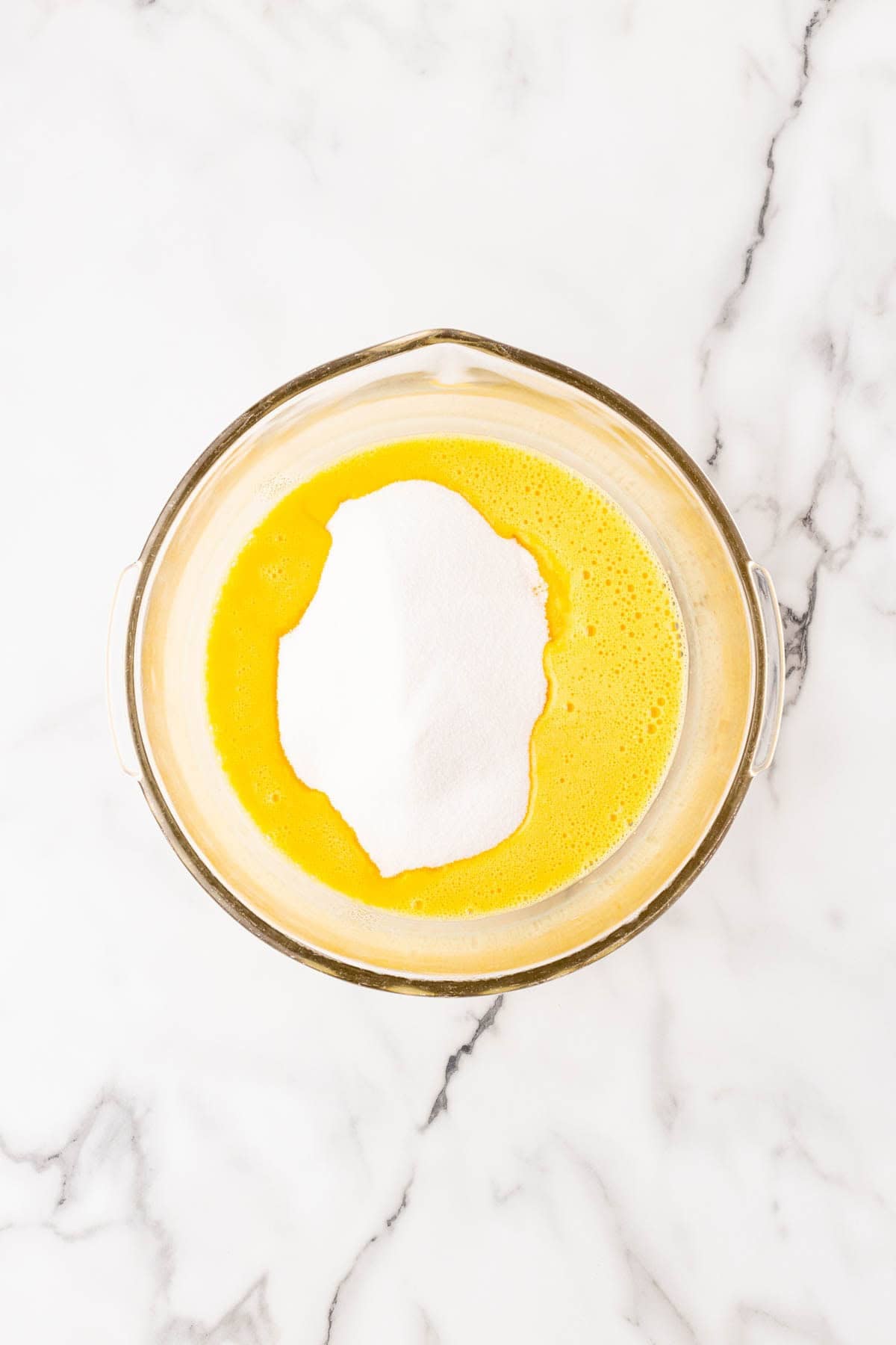 Granulated sugar added to beaten eggs in a glass mixing bowl on a marble countertop