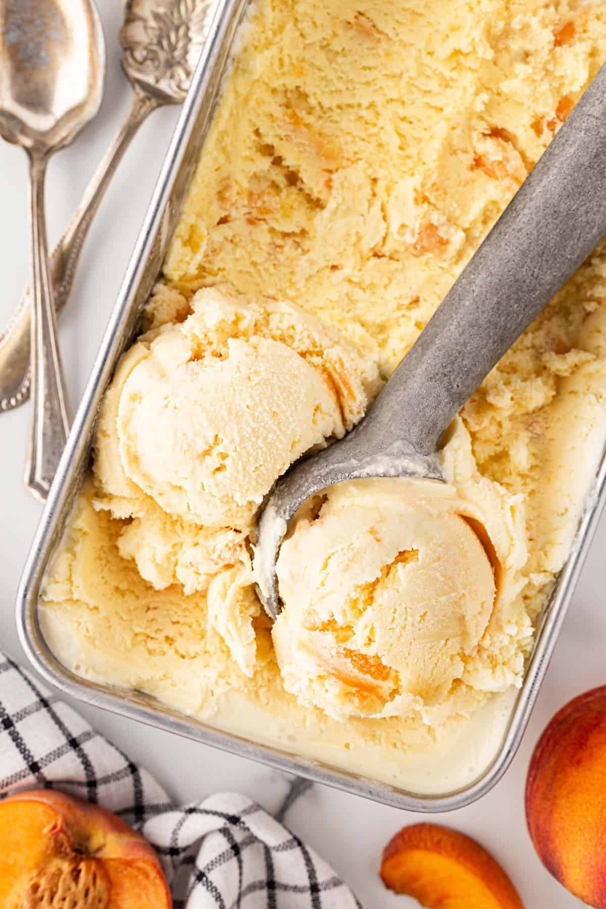 Overhead shot of homemade peach ice cream in a loaf pan with scoops, fresh peach slices, and vintage silver spoons
