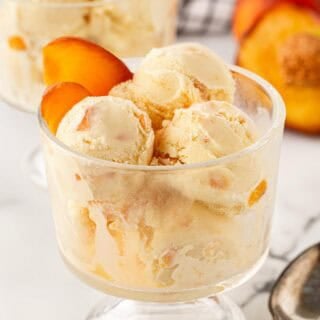 Scoops of peach ice cream served in a glass dessert dish with fresh peach slices, additional ice cream in the background, and vintage silver spoons on a marble surface