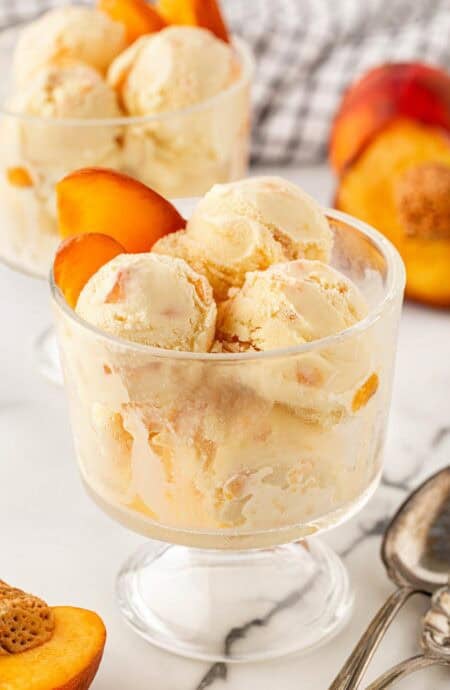 Scoops of peach ice cream served in a glass dessert dish with fresh peach slices, additional ice cream in the background, and vintage silver spoons on a marble surface