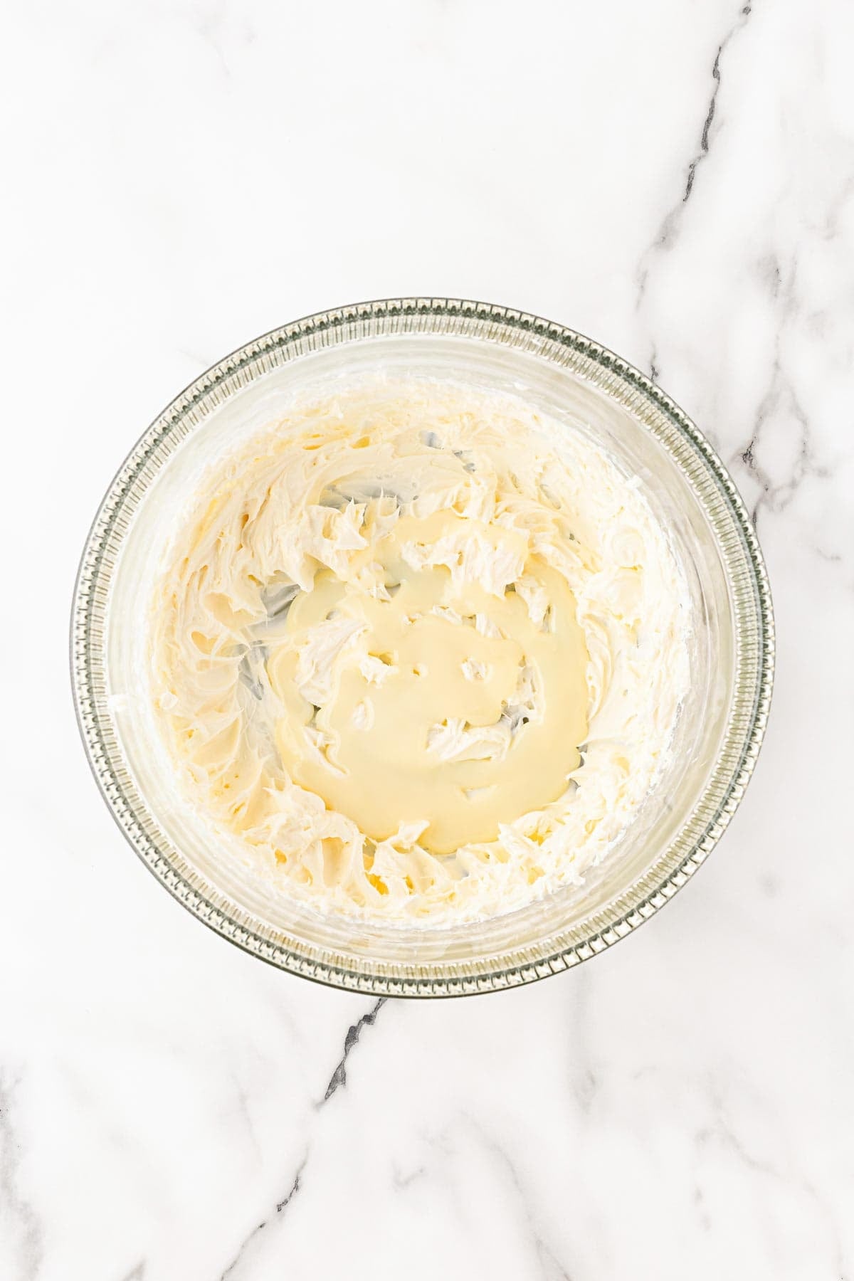 Sweetened condensed milk added to whipped cream cheese in a glass mixing bowl on a white marble background