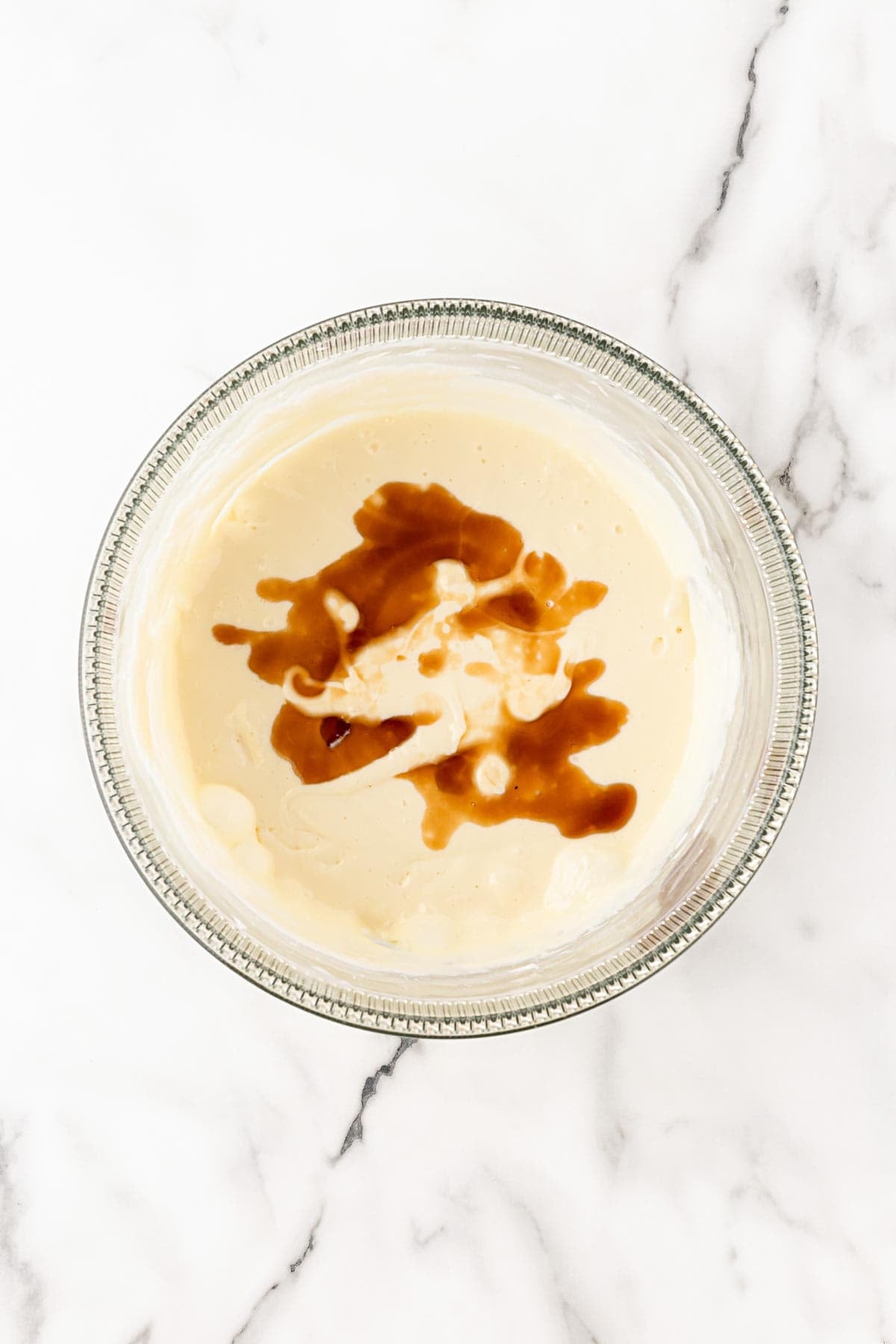 Vanilla extract poured into the cream cheese mixture in a glass mixing bowl on a white marble background