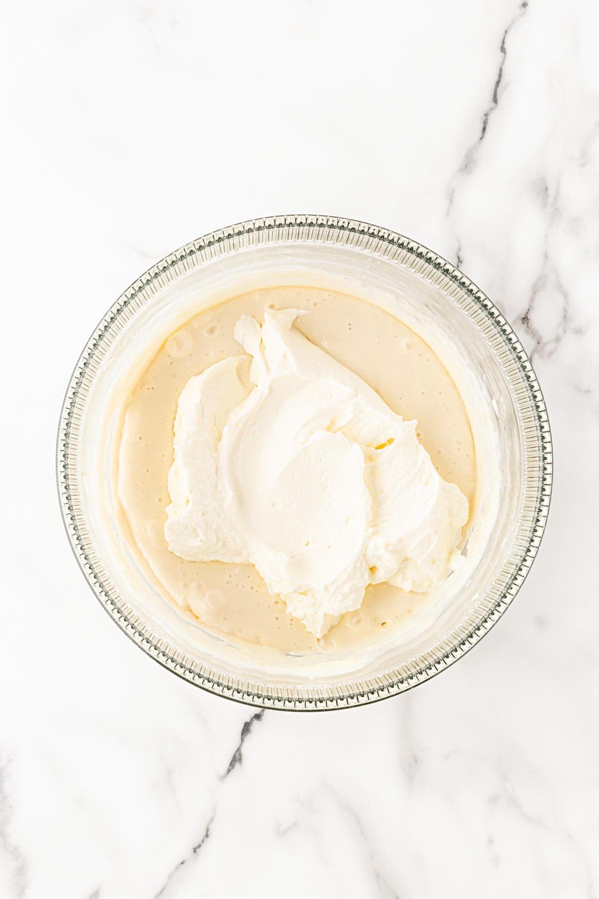 Whipped cream added to the cream cheese mixture in a glass mixing bowl on a white marble background