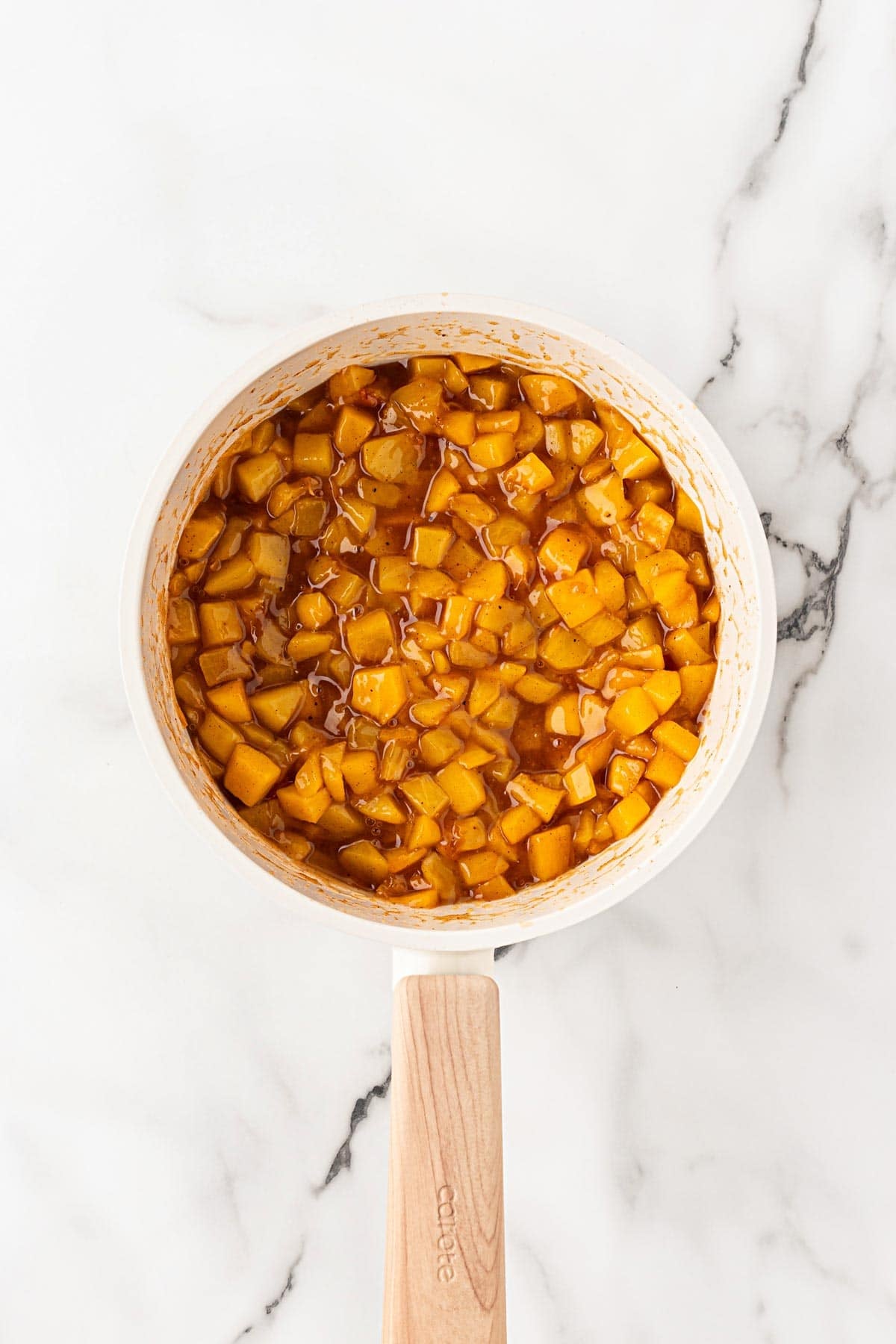 Diced peaches simmering in a spiced syrup in a light saucepan on a white marble background