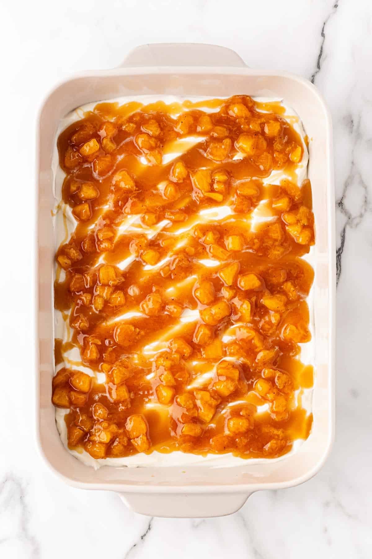 Caramel and spiced peach mixture spooned over the creamy layer in a baking dish on a white marble background