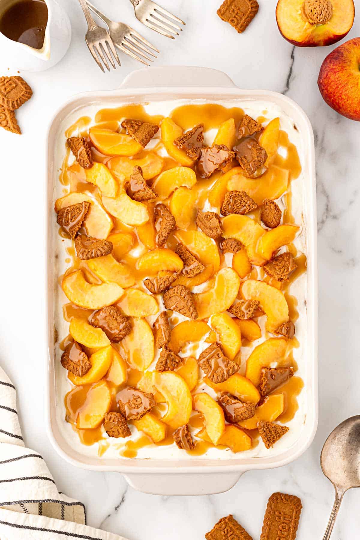 Peach pudding topped with fresh peach slices, Biscoff cookie pieces, and caramel drizzle, surrounded by peaches and utensils