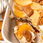 Spoon scooping a serving of peach pudding with layers of cream, peaches, Biscoff cookies, and caramel from the baking dish