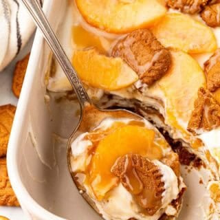Spoon scooping a serving of peach pudding with layers of cream, peaches, Biscoff cookies, and caramel from the baking dish