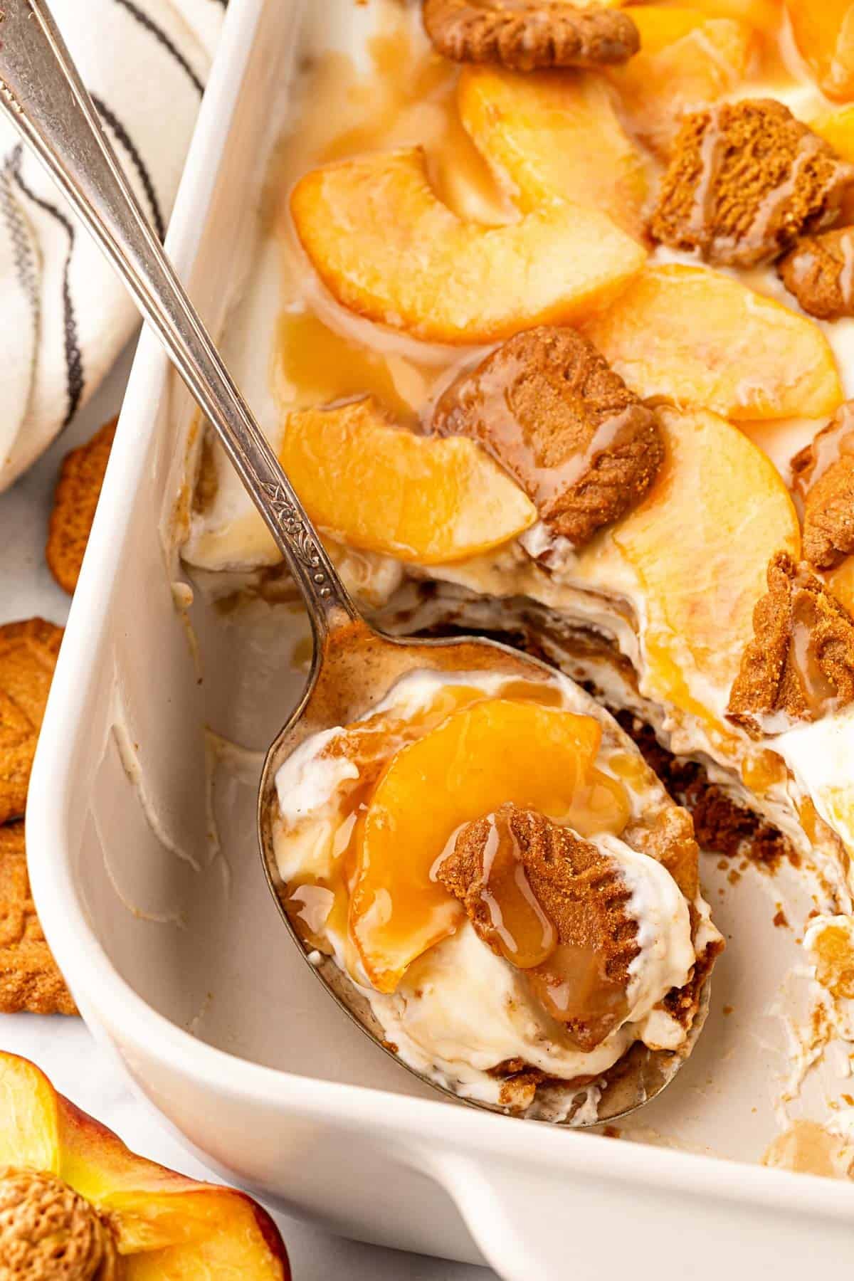 Spoon scooping a serving of peach pudding with layers of cream, peaches, Biscoff cookies, and caramel from the baking dish