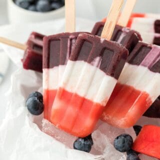 Red, white, and blue popsicles served over ice with fresh blueberries and watermelon slices in the background