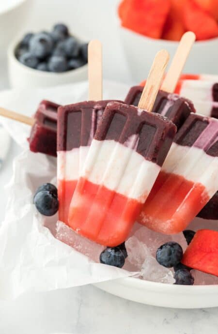 Red, white, and blue popsicles served over ice with fresh blueberries and watermelon slices in the background