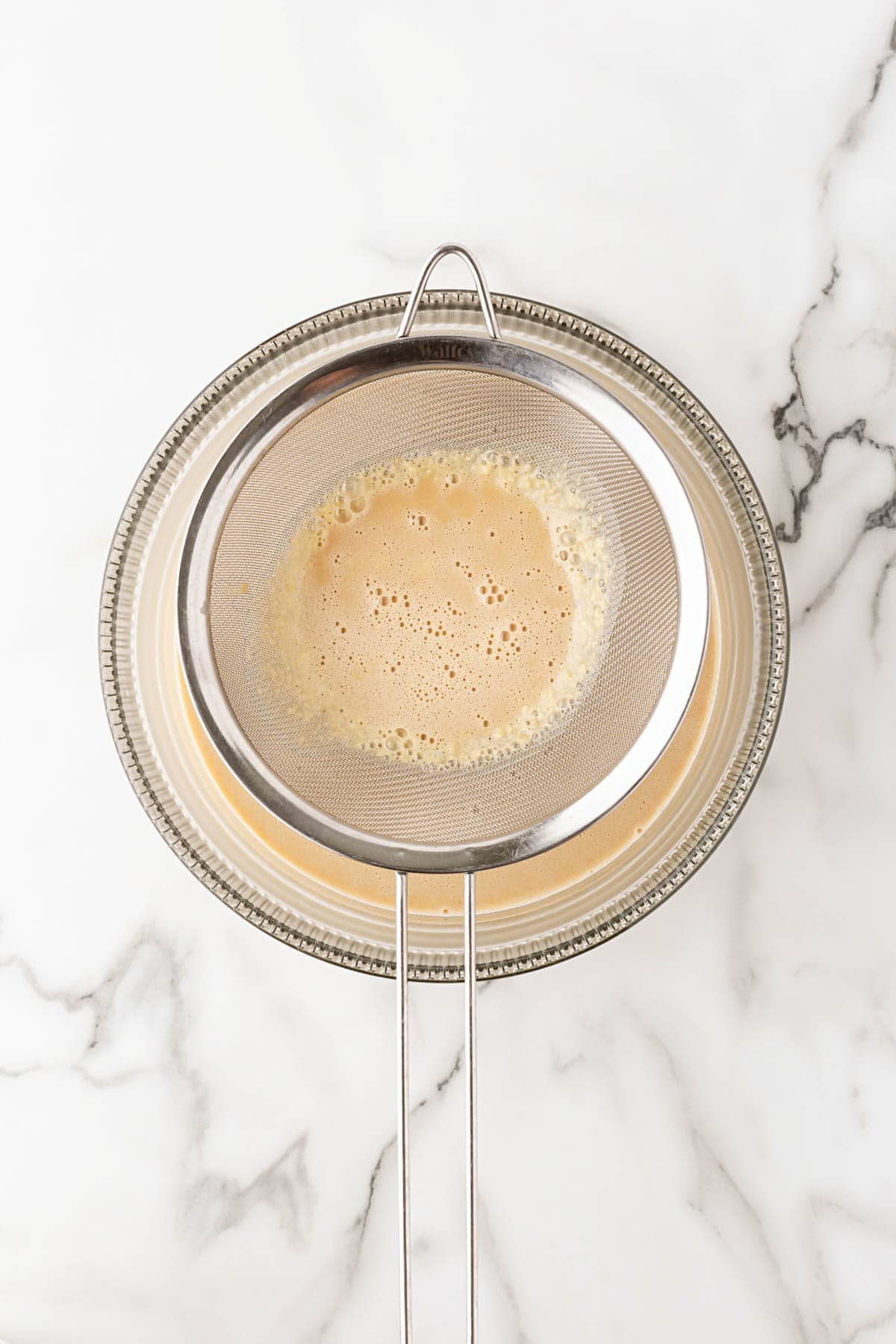 Biscoff ice cream custard strained through a fine-mesh sieve into a glass bowl on a white marble countertop