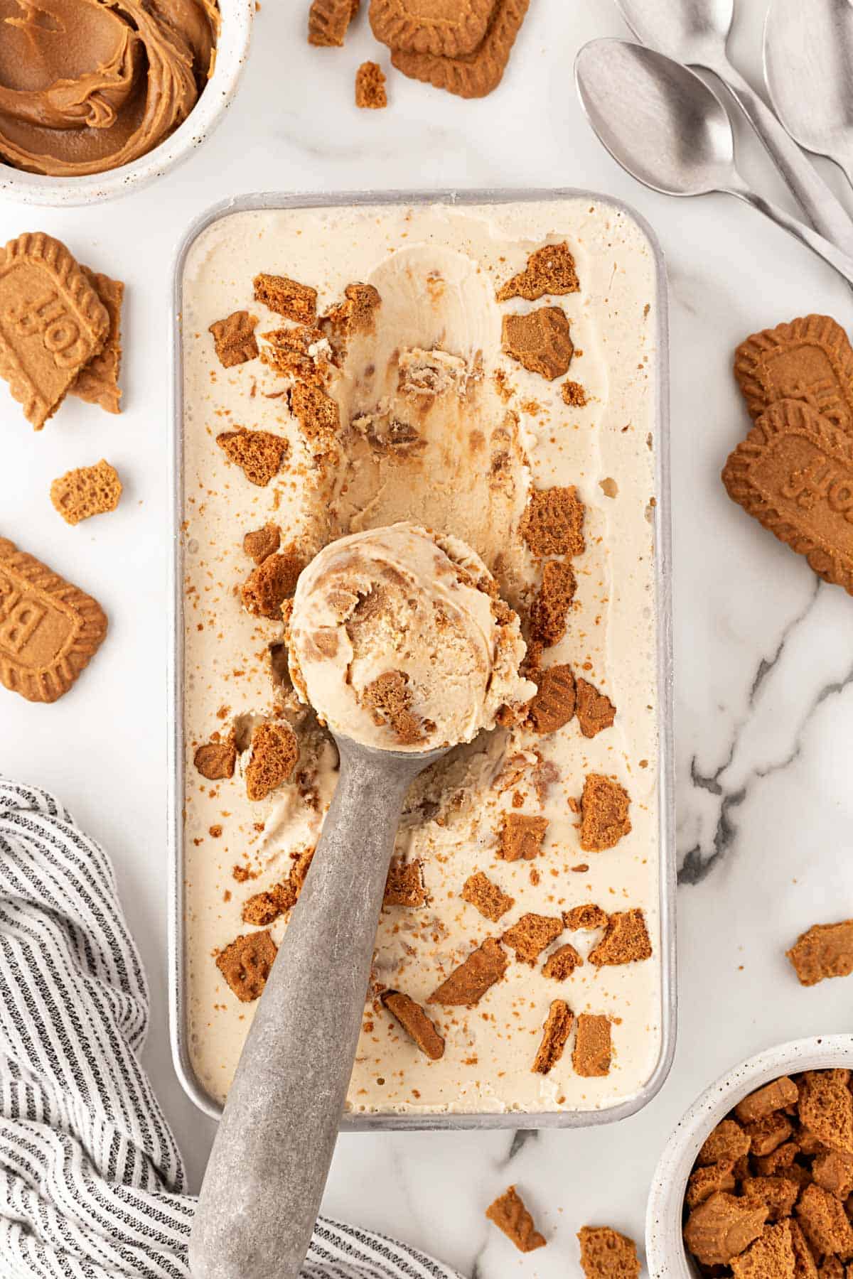 Scoop of Biscoff ice cream lifted from a pan topped with crushed cookies, with cookie butter and cookies surrounding it