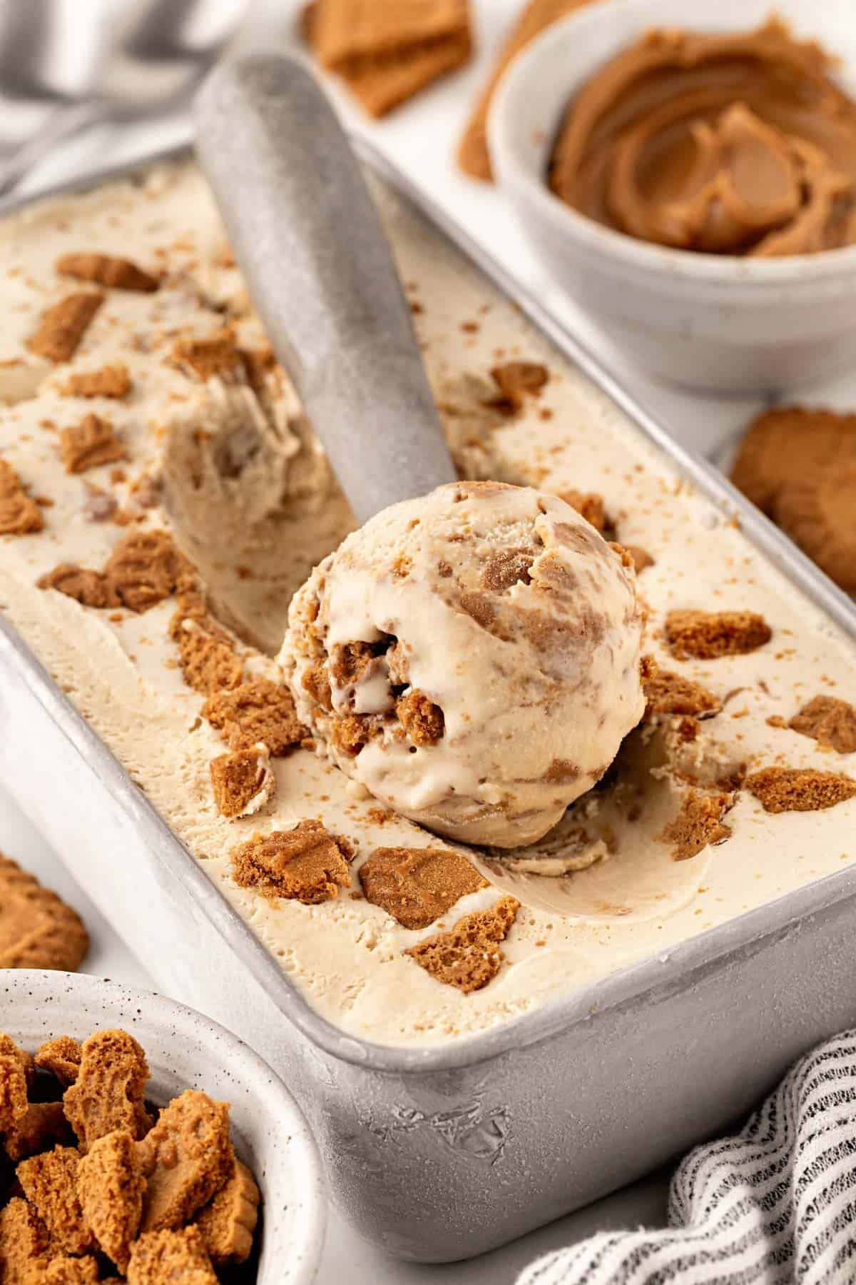Scoop of Biscoff ice cream with cookie pieces lifted from a metal pan, with Biscoff spread and cookies in the background