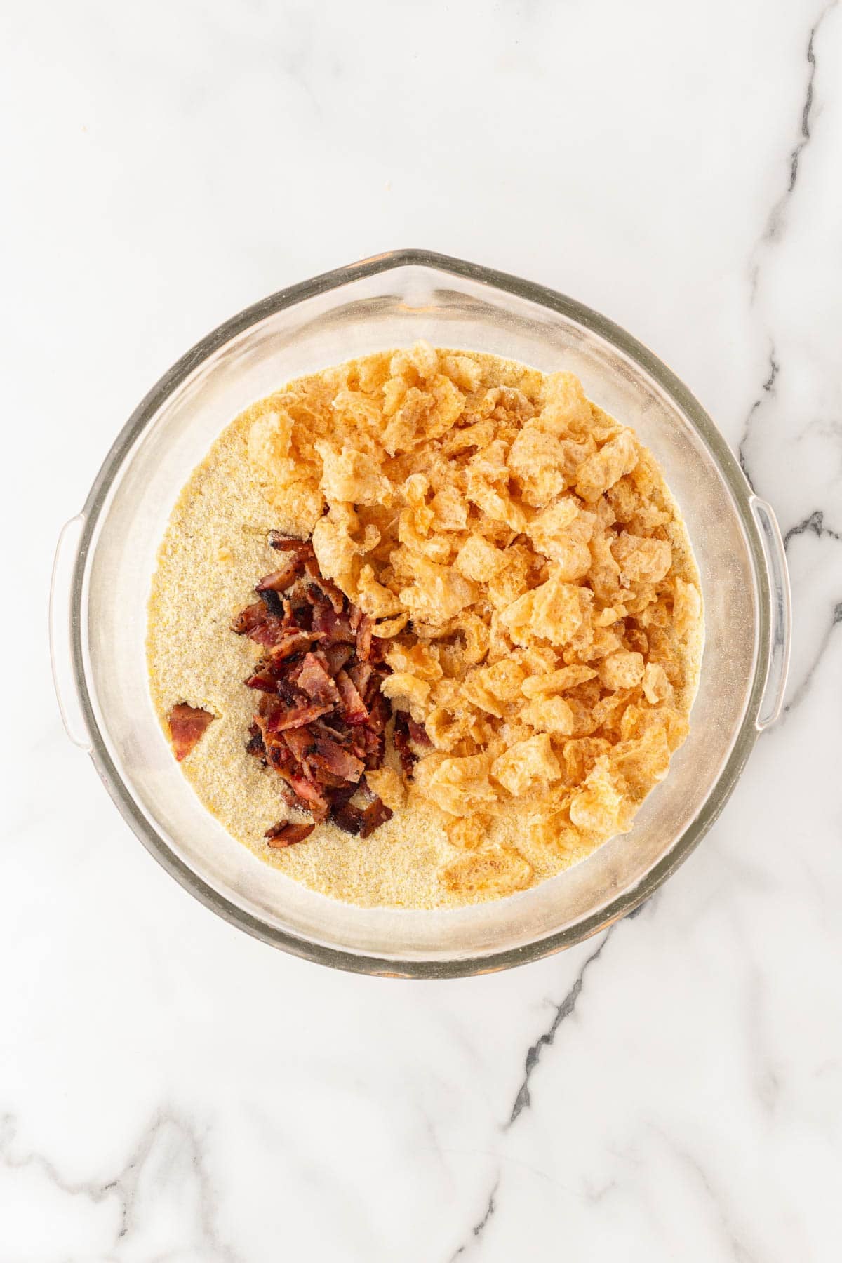 Glass mixing bowl with cornmeal, chopped bacon, and pork cracklings on a white marble countertop