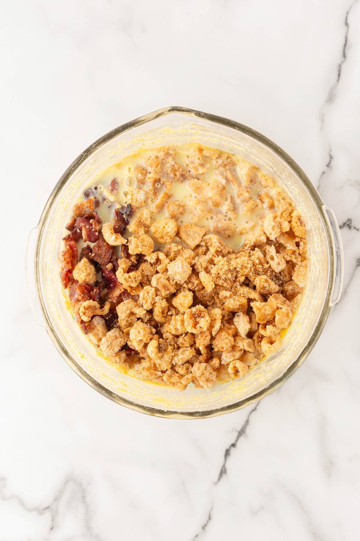 Glass mixing bowl with cornmeal, chopped bacon, pork cracklings, and wet ingredients added on a white marble countertop