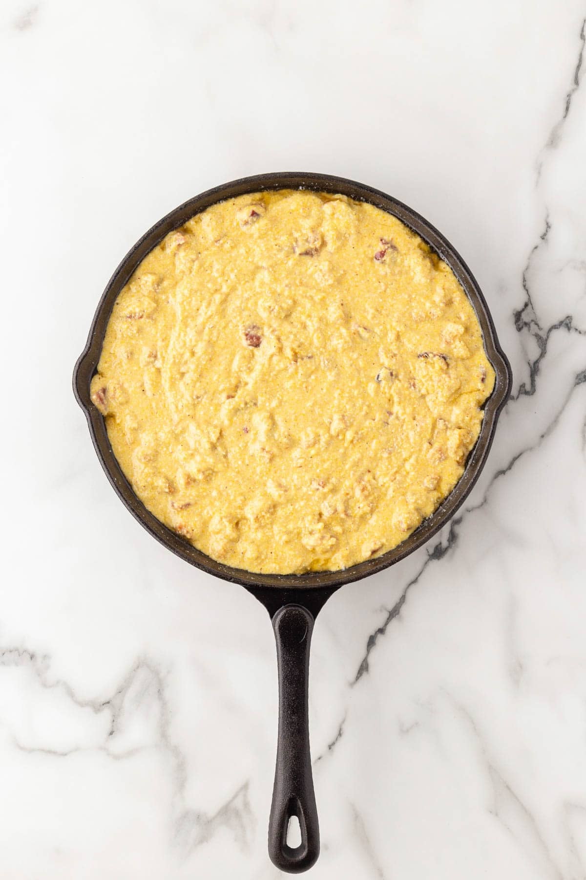 Crackling cornbread batter spread evenly in a buttered cast iron skillet on a white marble countertop