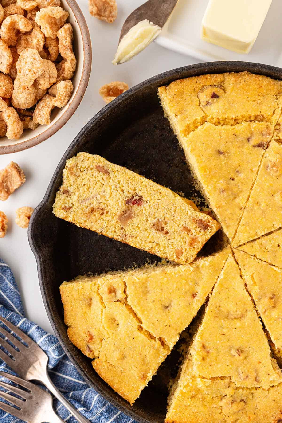 Overhead shot of sliced crackling bread in a cast iron skillet with butter, pork cracklings, and blue striped kitchen towel